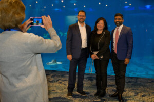 Three people in front of water tank from the georiga aquarium in the ocean's ballroom and someone taking a picture of three people with an iphone