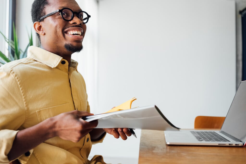 Man Smiling With Yellow Shirt At Computer Holding Paper
