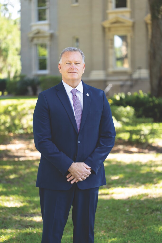 A white male with gray hair in a blue suit and purple tie stands in front of a building with green gras and shrubs.