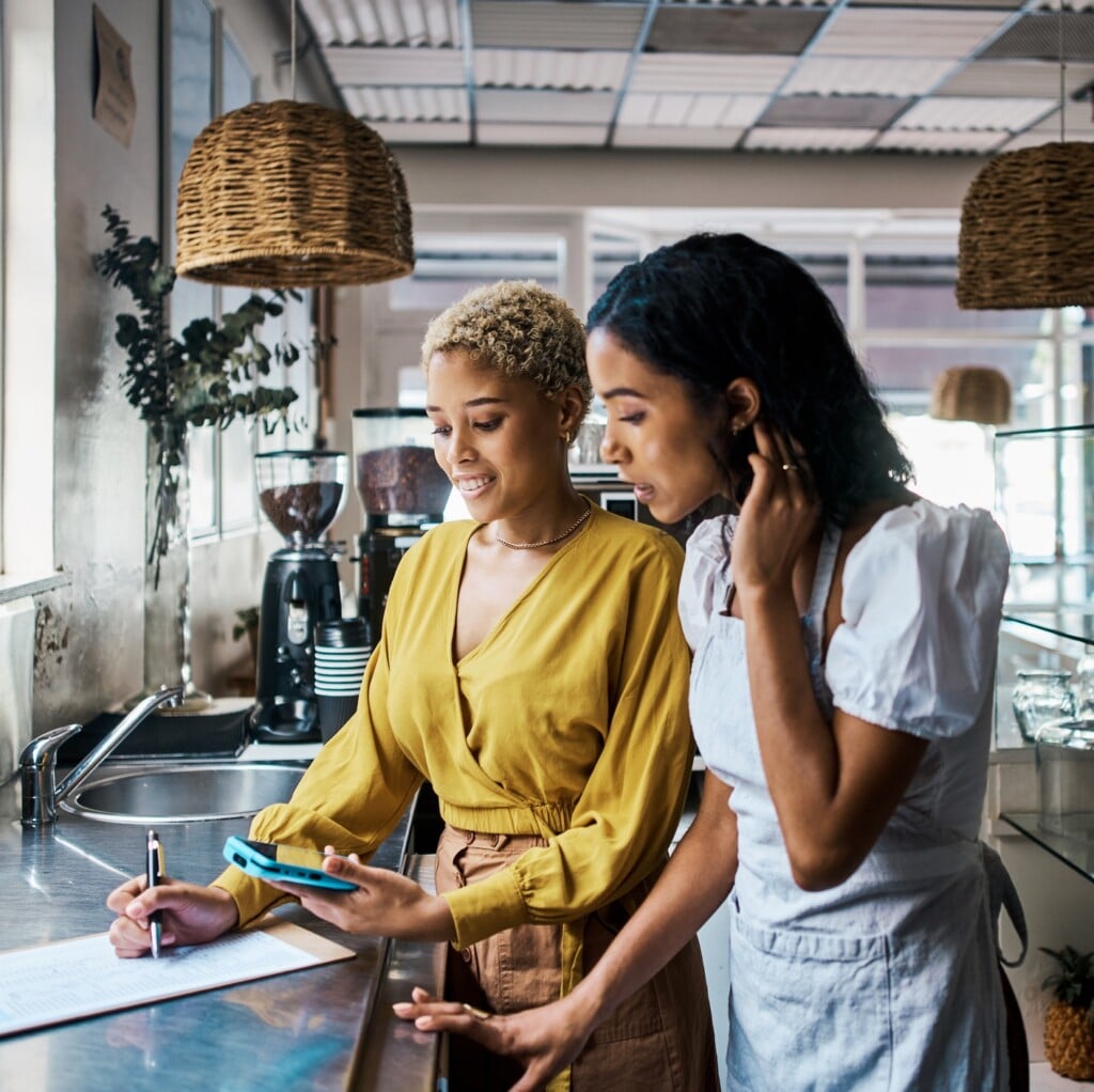 Small Business Owners Working Together And Writing Down Orders In A Coffee Shop. Female Entrepreneurs In Partnership Collaborating, Brainstorming And Planning Sales For Their Cafe in Georgia