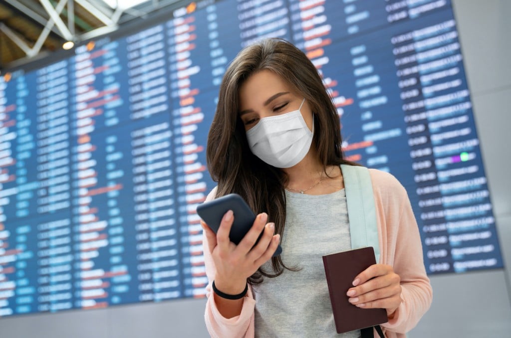 Female Traveler Wearing A Facemask At The Airport While Texting On Her Phone