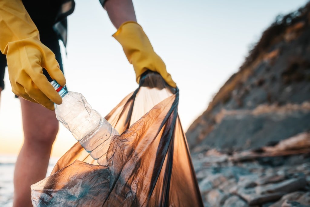 A Woman Volunteer Puts A Plastic Bottle In A Polyethylene Bag. Close Up Of Hands. In The Background Wild Beach And Ocean. The Concept Of Environmental Conservation And Clean Up Of Coast