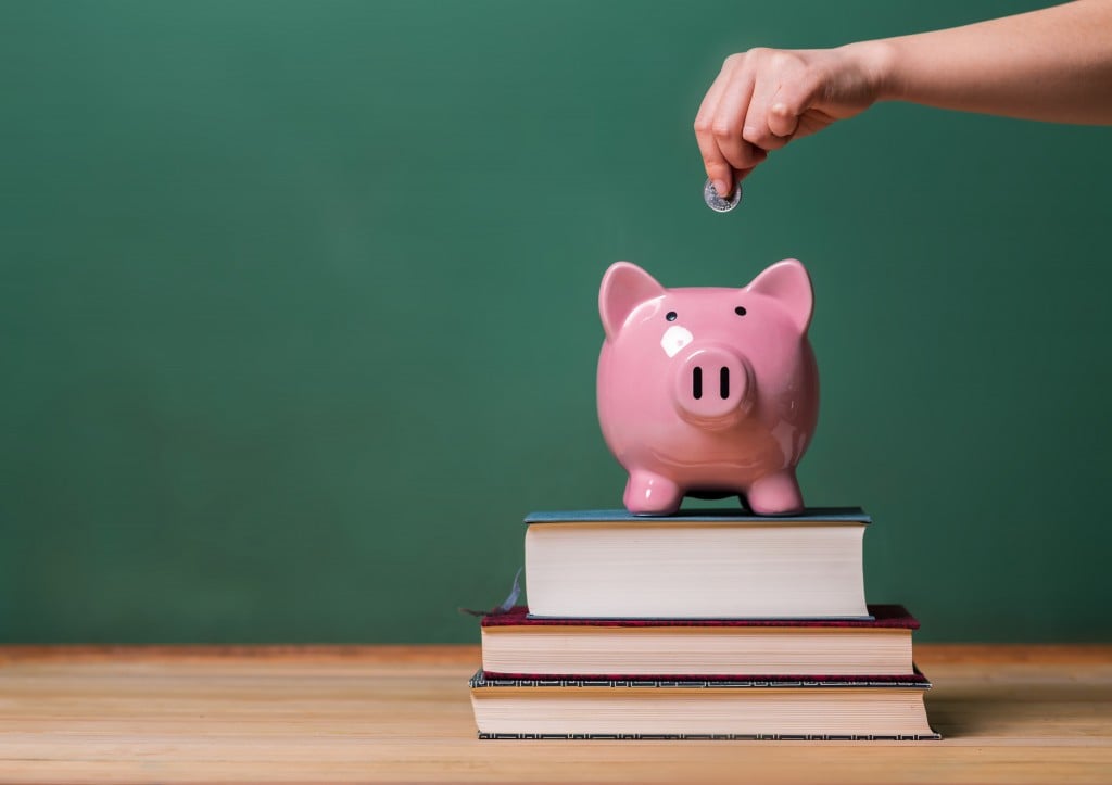 Person Depositing Money In A Piggy Bank On Top Of Books With Chalkboard