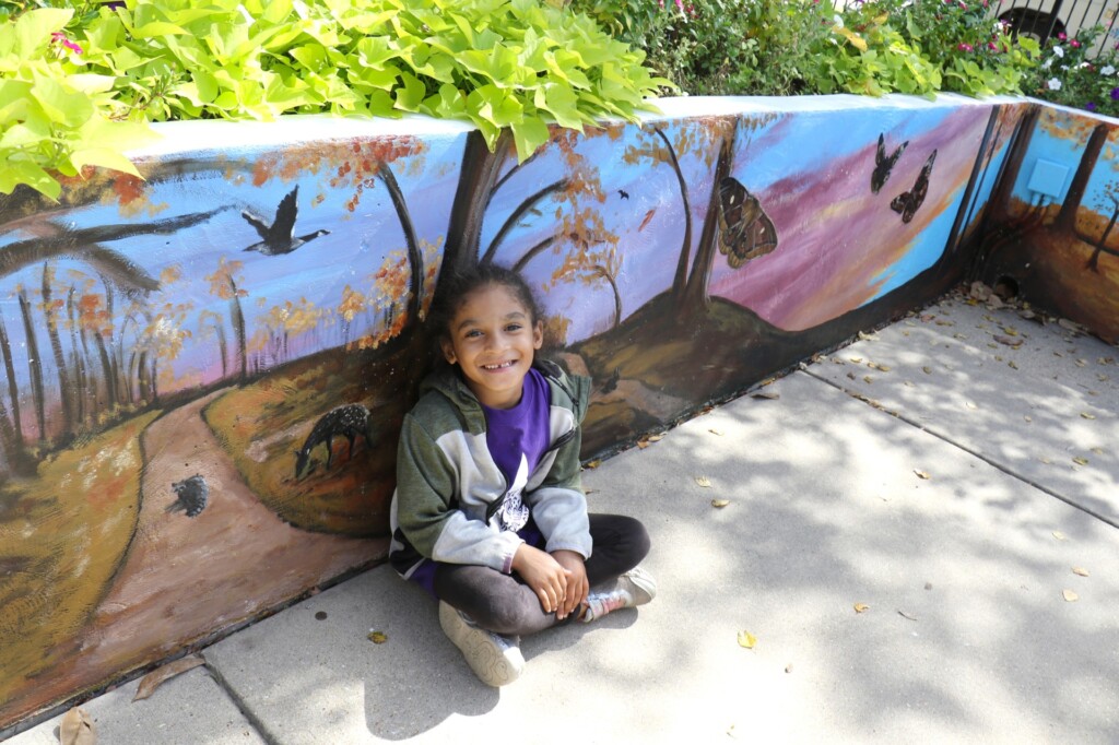 Kindergartner Zolah Jones of Margaret Peacock’s class sits beside an autumn scene from the new mural in the sensory garden of the Poplar Bluff Municipal Library. (Source: Poplar Bluff R-I School District)