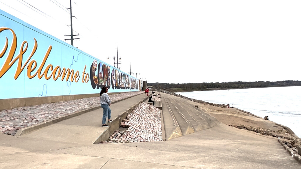 People walk on the sidewalk in front of a wall with a mural for Cape Girardeau. The river is seen to the right of the photo.