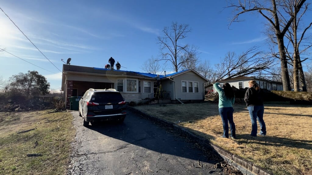 Two women stand on the grass in front of a home as men stand on the rooftop, making repairs due to storm damage