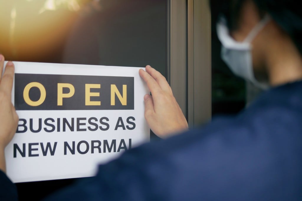 Reopening For Business Adapt To New Normal In The Novel Coronavirus Covid 19 Pandemic. Rear View Of Business Owner Wearing Medical Mask Placing Open Sign "open Business As New Normal" On Front Door.