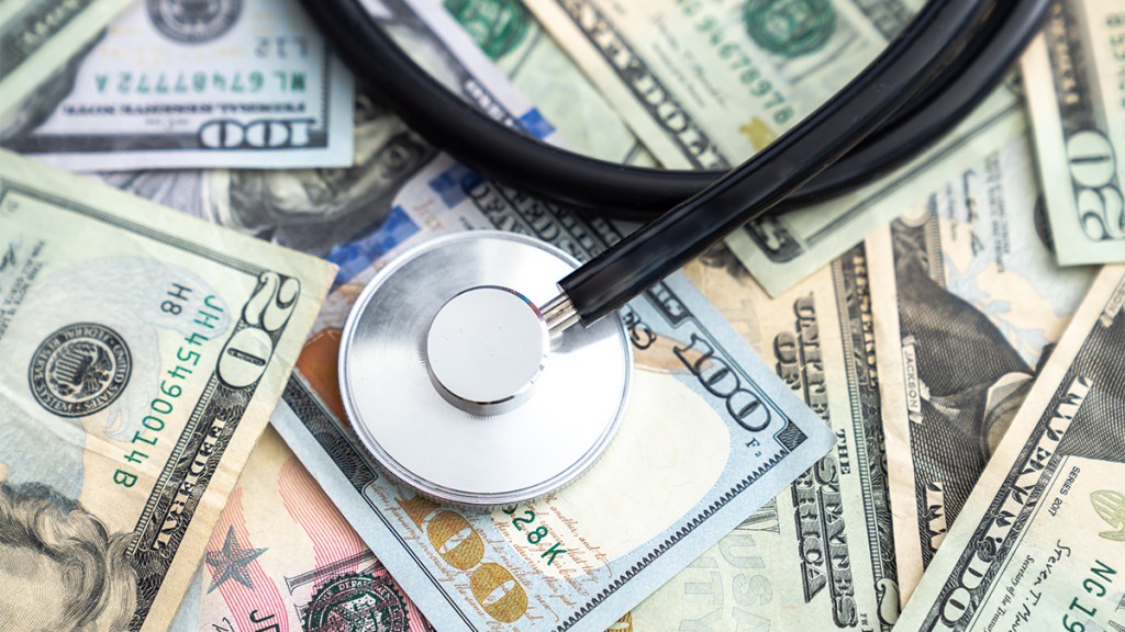 Doctor Or Nurse Stethoscope Medical Device Or Equipment With Metal Parts And Black Tubing Laying On A Pile Of United States Currency In Large Bills Or Cash Covering The Entire Surface.