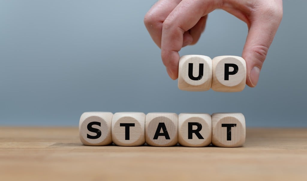 Cubes Form The Words "start Up" While To Fingers Lift The Letters "up" In The Air. Cubes Are On A Wooden Table In Front A Grey Background.