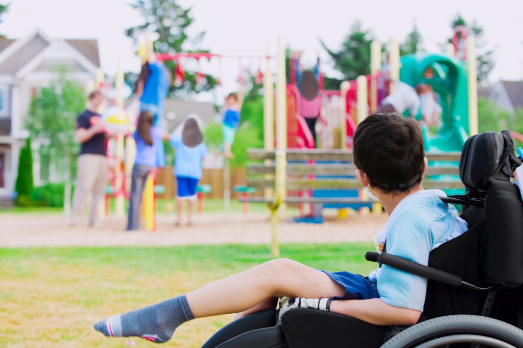 Disabled Little Boy In Wheelchair Watching Children Play On Playground