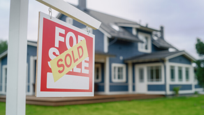 Close Up Of A House Sold Sign On A Lawn In Front Of A Big Modern House With Traditional Architecture. Housing Market Concept With Residential Property In The Countryside.