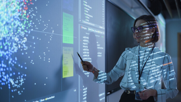 Young Female Teacher Giving A Data Science Presentation In A Dark Auditorium With Projecting Slideshow With Artificial Intelligence Neural Network Architecture. Business Startup And Education Concept