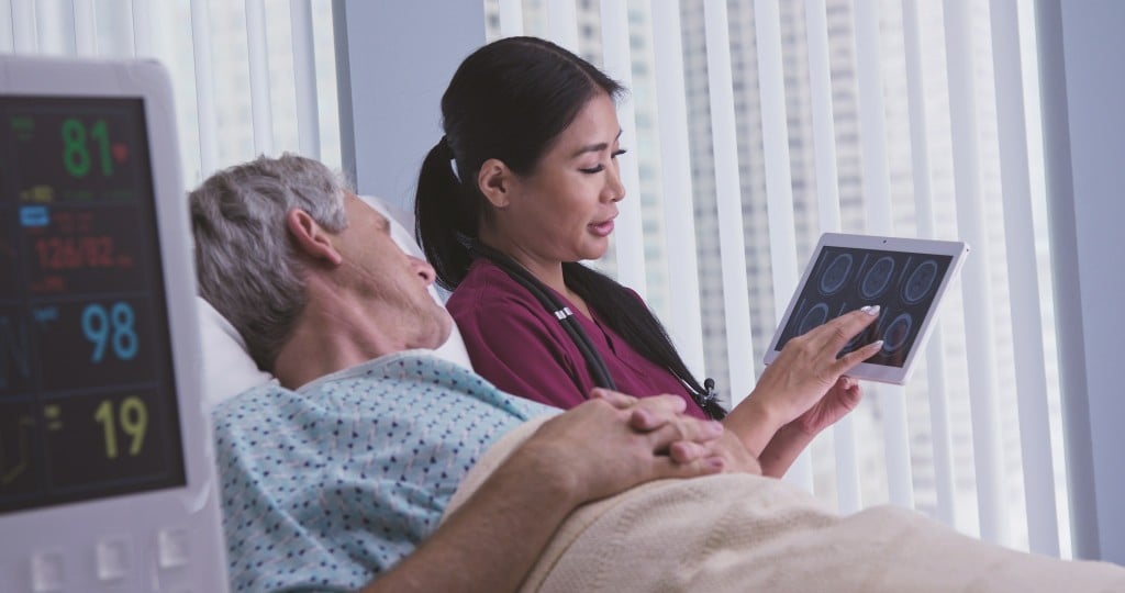 Doctor Sharing Test Results On Tablet Computer With Patient In Hospital Bed