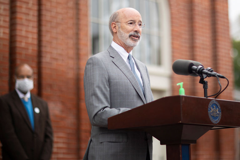 Govenor Wolf Speaking At A Podium Outside Of A Brick Building