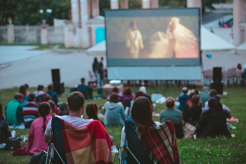 Couple Sitting In Camp Chairs In Front Of Movie Screen At Open Air Cinema