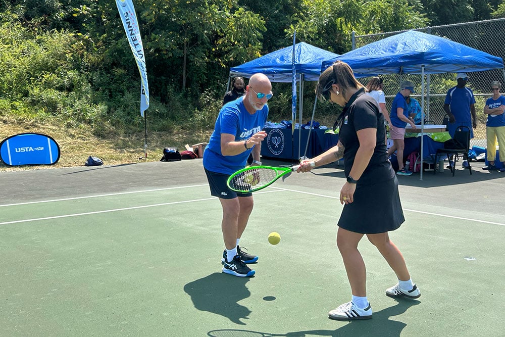 A man and woman stand on a tennis court. The woman, on the right, is holding a tennis racket in her right hand. She's bouncing a tennis ball that is mid-air at knee-length.