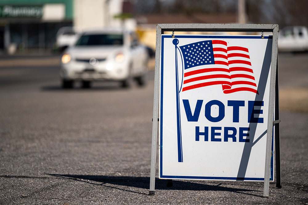 A sign on the ground that reads "Vote Here" below a graphic of the American Flag.