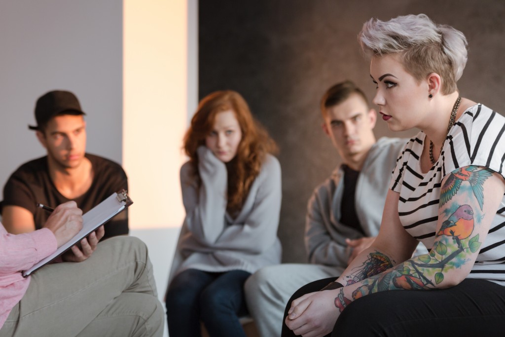 Tattooed Rebel Teenage Girl Sitting In Front Of A Therapist Who Is Taking Notes During A Group Psychotherapy Meeting. Other Young People In The Blurred Background.