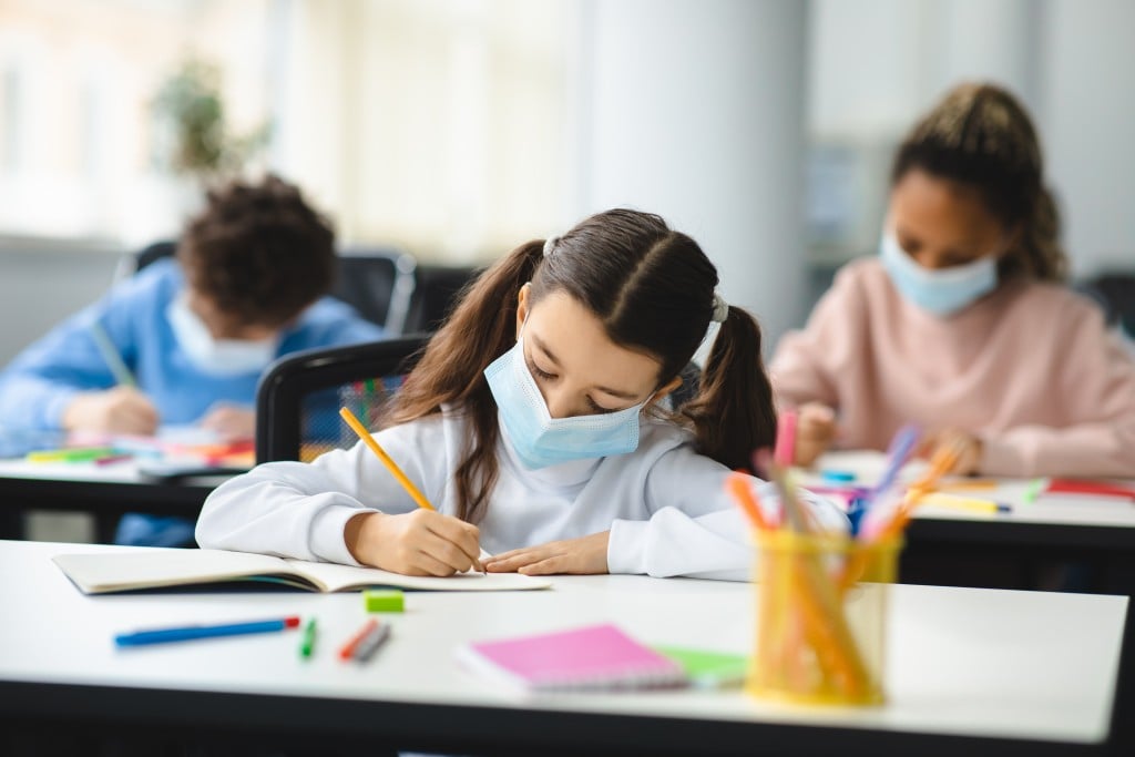 Girl In Mask Sitting At Desk Writing In Classroom