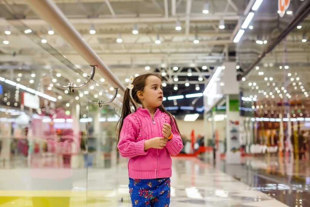 Little Girl Alone In Shopping Mall Center, Looking For Her Mother. The Child Was Lost In The Big Store.