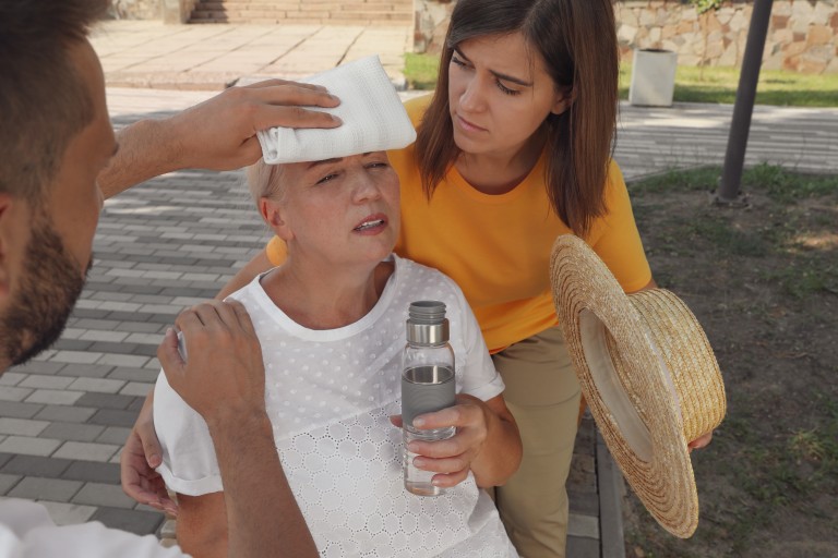 People Helping Mature Woman On City Street. Suffering From Heat Stroke