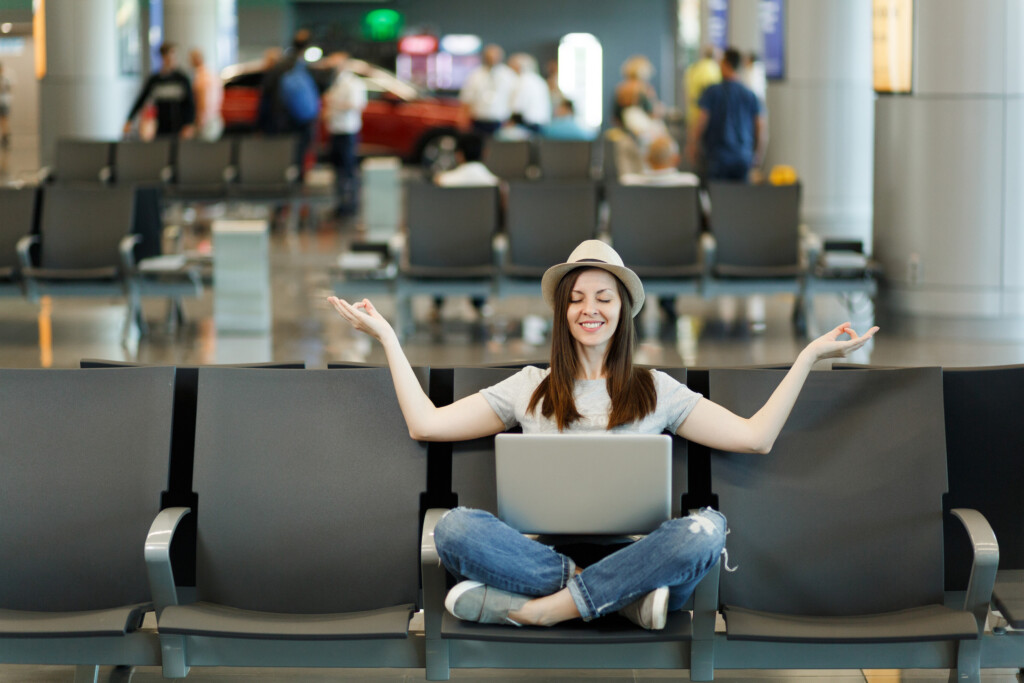 Young Smiling Traveler Tourist Woman With Laptop Sitting With Crossed Legs, Meditate, Spread Hands, Waiting In Lobby Hall At Airport. Passenger Traveling Abroad On Weekend Getaway. Air Flight Concept.