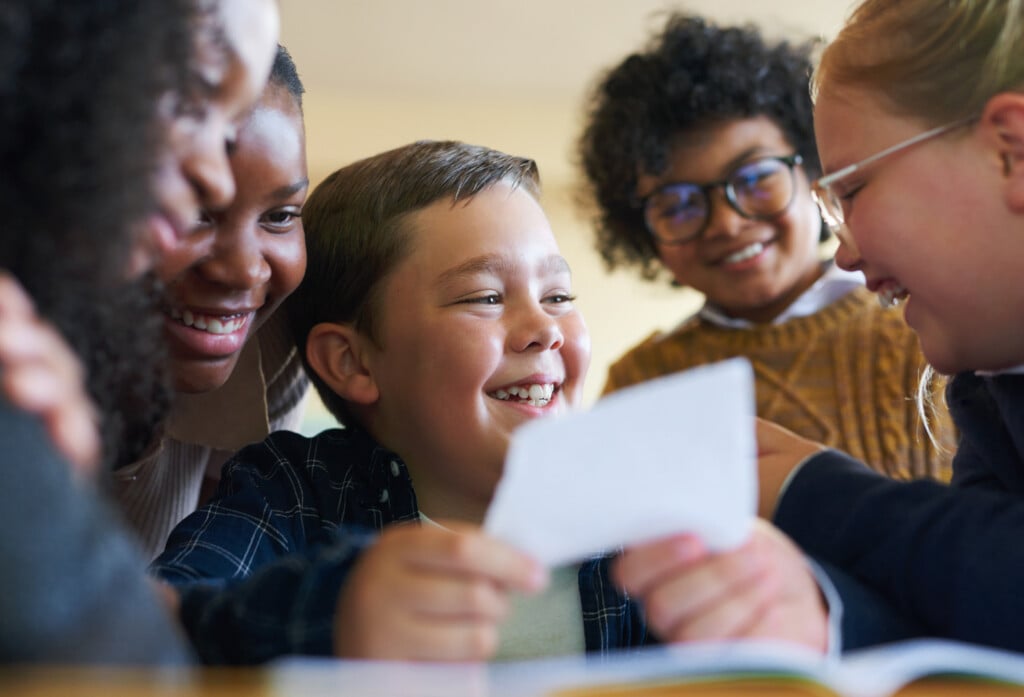 What Do I Say To This Note. A Diverse Group Of Children Huddled Together In Their School Classroom And Laughing While Reading A Note.