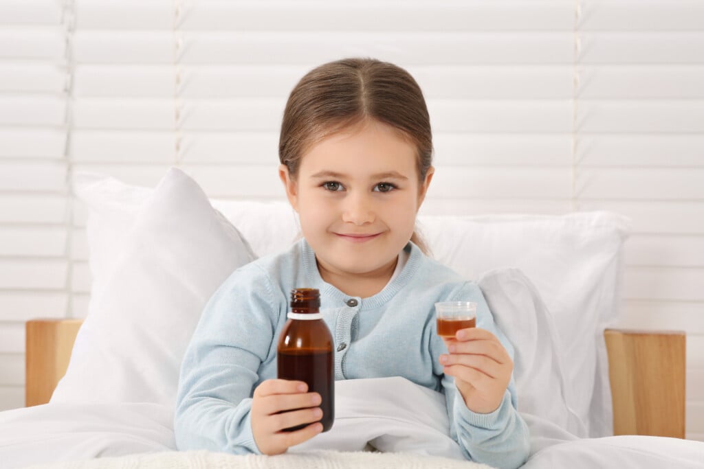 Cute Girl Holding Bottle And Measuring Cup With Cough Syrup In B