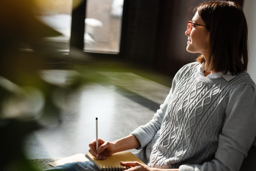 White Young Woman Writing Down Notes While Sitting On Floor At Home