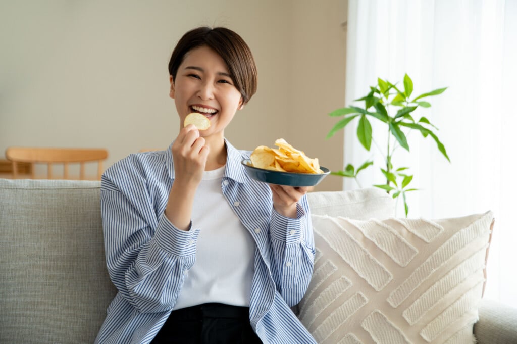 Young Woman Eating Potato Chips And Smiling While Sitting On Sofa At Home