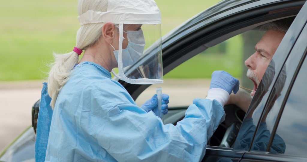 Dressed In Full Protective Gear A Healthcare Worker Collects A Sample From A Mature Man Sitting Inside His Car As Part Of The Operations Of A Coronavirus Mobile Testing Unit...