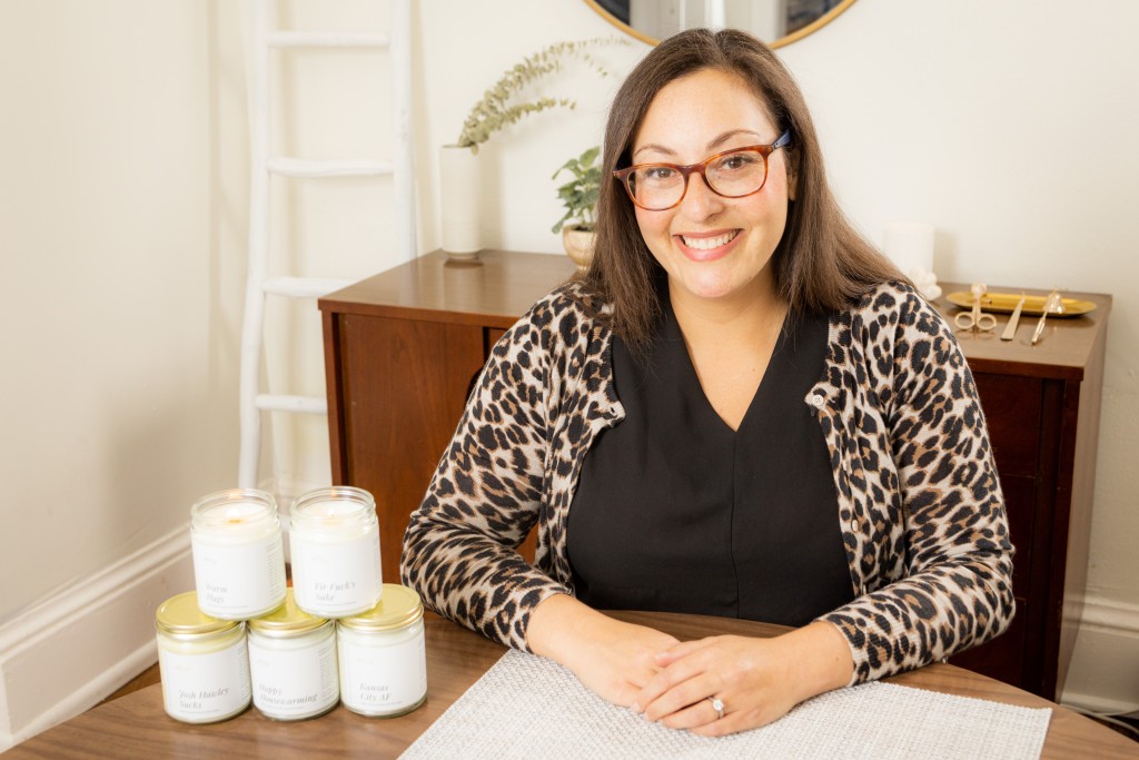 Birdie Hansen, of Effing Candle Co., sits with her arms on the table in front of her. A stack of five candles with white labels and gold lids sits to her right. Behind her are some wick trimming tools on a wooden credenza.