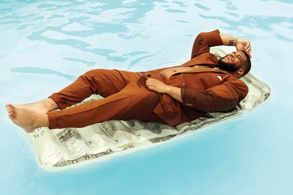 Steven G. wearing a brown suit in a pool, floating atop a floatie shaped like a dollar bill