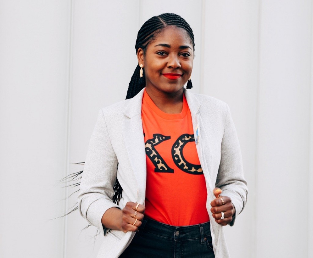Chelsey from KC Black Owned stands front and center in the photo, holding her blazer in a powerful stance. She is wearing black jeans, a white blazer, and a red shirt with a cheetah-print "KC" logo.
