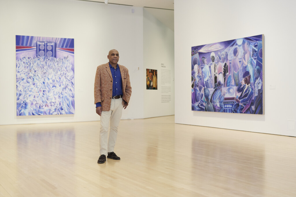 Denzil Forrester stands in front of some of his paintings in his exhibition "Denzil Forrester: Duppy Conquerer."