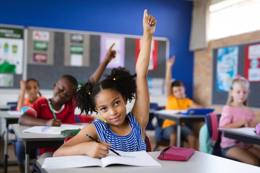 girl in classroom raising hand, for article on questions to ask when choosing a school