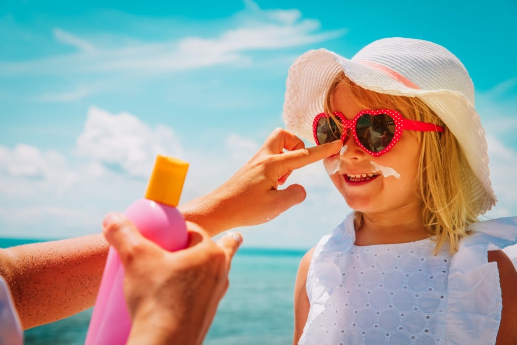 young girl in sunhat and glasses has sunscreen applied to her face