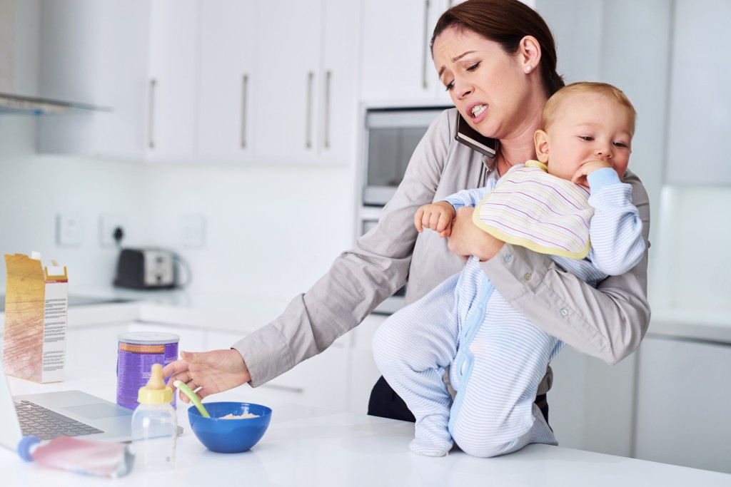 mom looking stressed holding baby talking on phone and making breakfast, for article on time management for moms