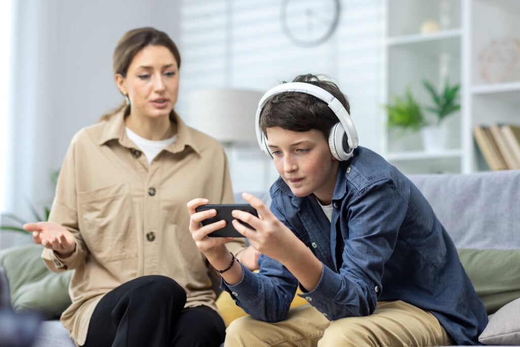 mom trying to talk to teenage son while he ignores her, wearing headphones and playing a video game