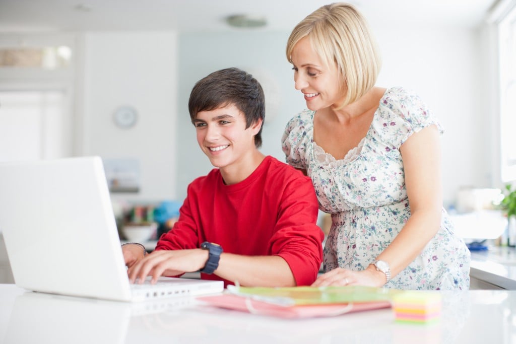 mom watching smiling teenage boy on laptop, for article on alternative education