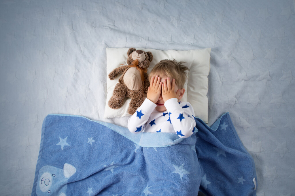young child in bed with hands over their face, for article on bedwetting