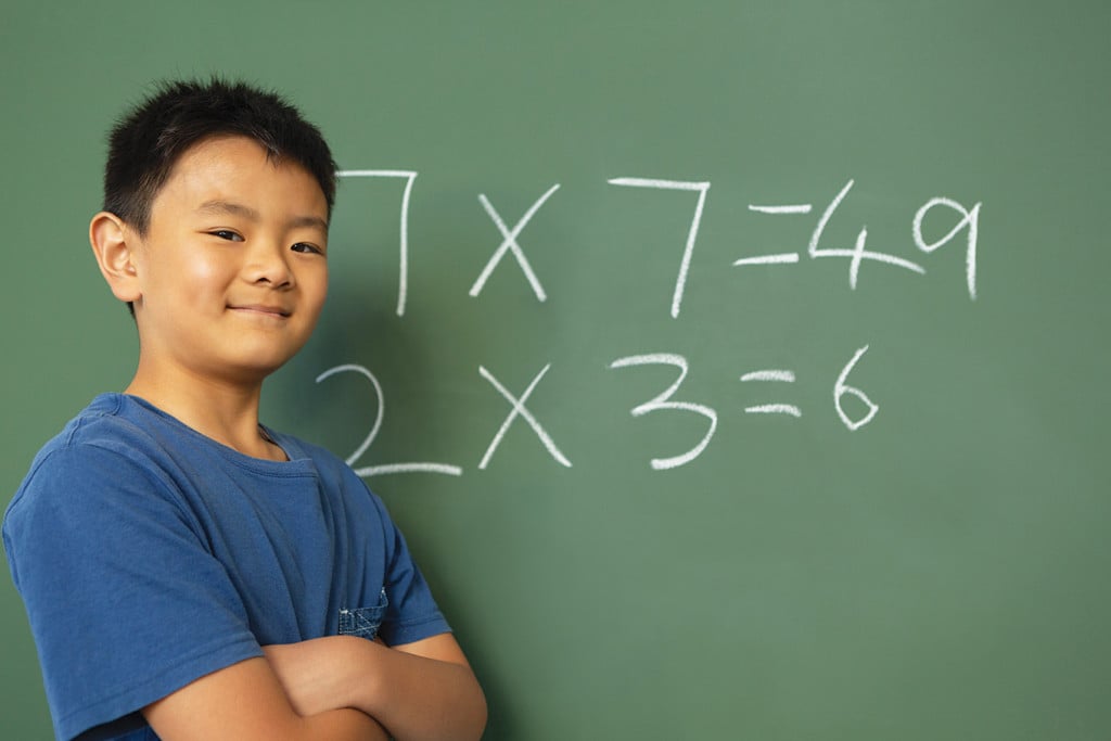 third grade boy stands in front of a chalkboard with math equations on it