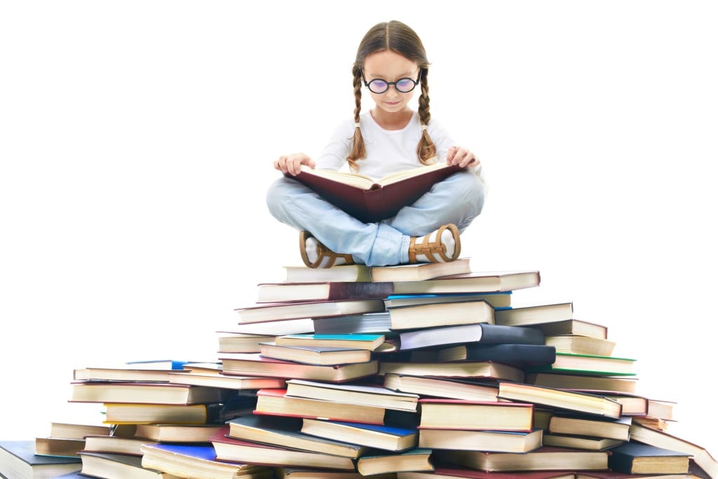 girl sitting on stack of books reading, for article on when to get library cards for kids