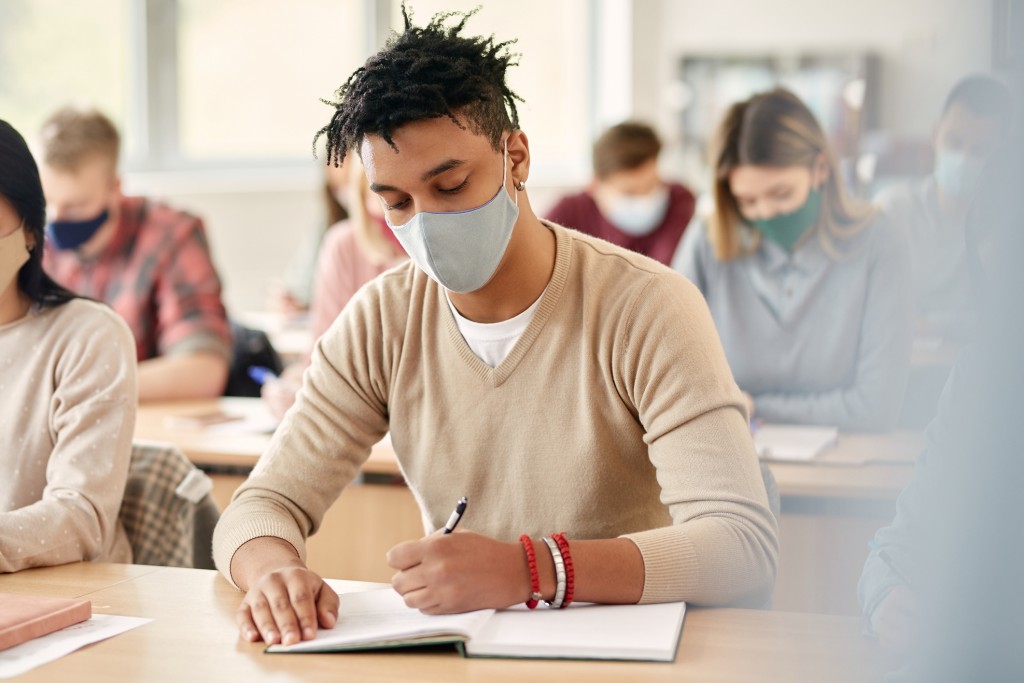 Black College Student With Face Mask Taking Notes During Lecture In The Classroom.