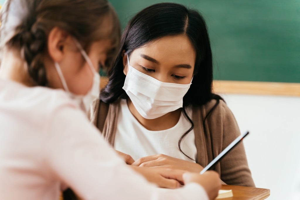 Close Up Of Asian Female Teacher Wearing A Face Mask In School Building Tutoring A Primary Student Girl. Elementary Pupil Is Writing And Learning In Classroom.