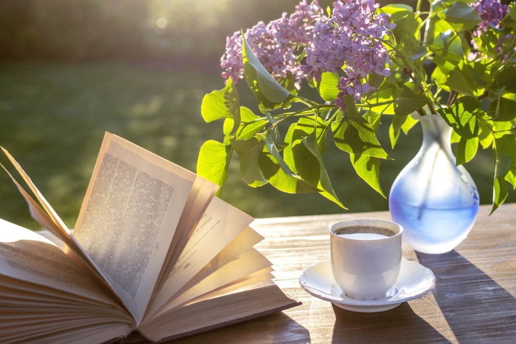 Little White Cup Of Espresso Coffee, Opened Book, Blue Semi Transparent Vase With Purple Lilac Flowers On Rustic Wooden Table In The Garden At Spring Morning After Sunrise Or At Evening Before Sunset