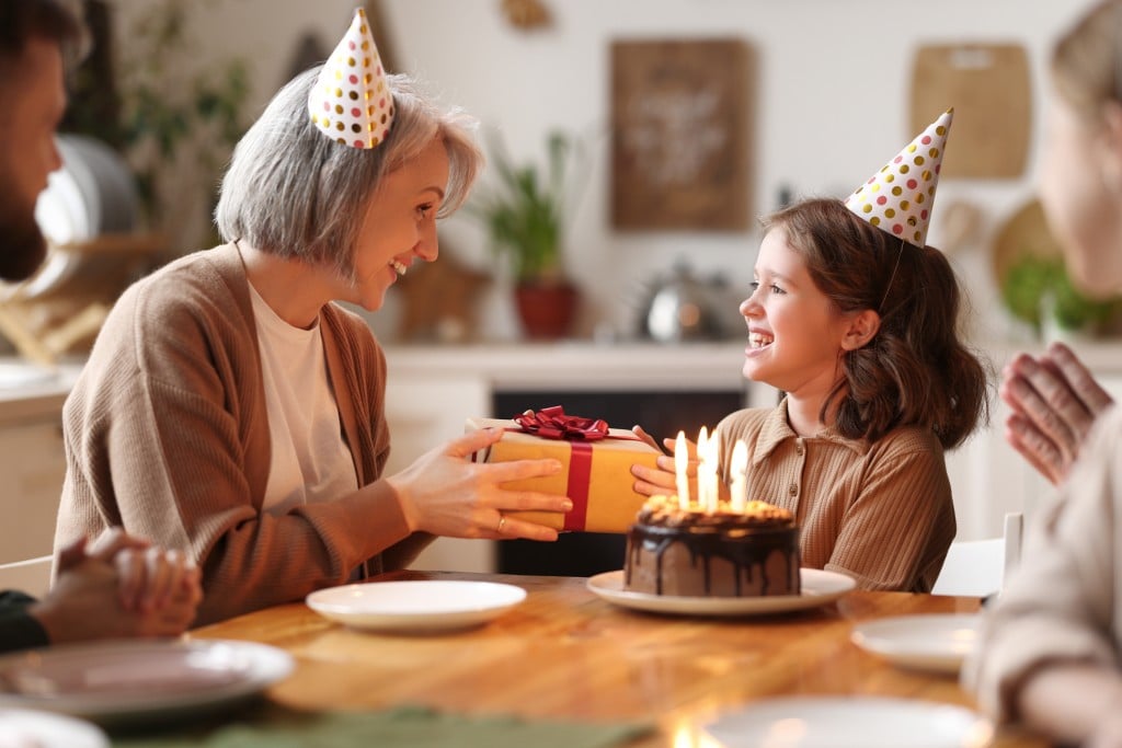 Loving Senior Woman Giving Gift To Excited Happy Granddaughter During Birthday Celebration With Family, for article on gift ideas for grandchildren