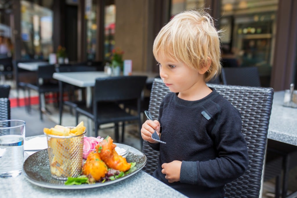 A young boy sits in front of a plate of food at a restaurant, outdoors, for article on dining out with b abies and toddlers