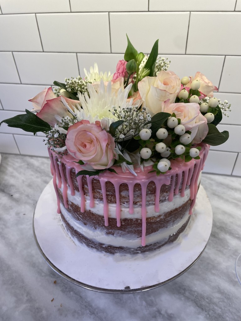 A naked cake with pink drip icing covered in light pink and white flowers made by ellen dale