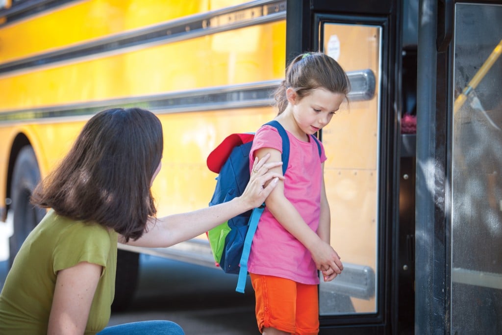 young girl with back-to-school anxiety by school bus with her mom